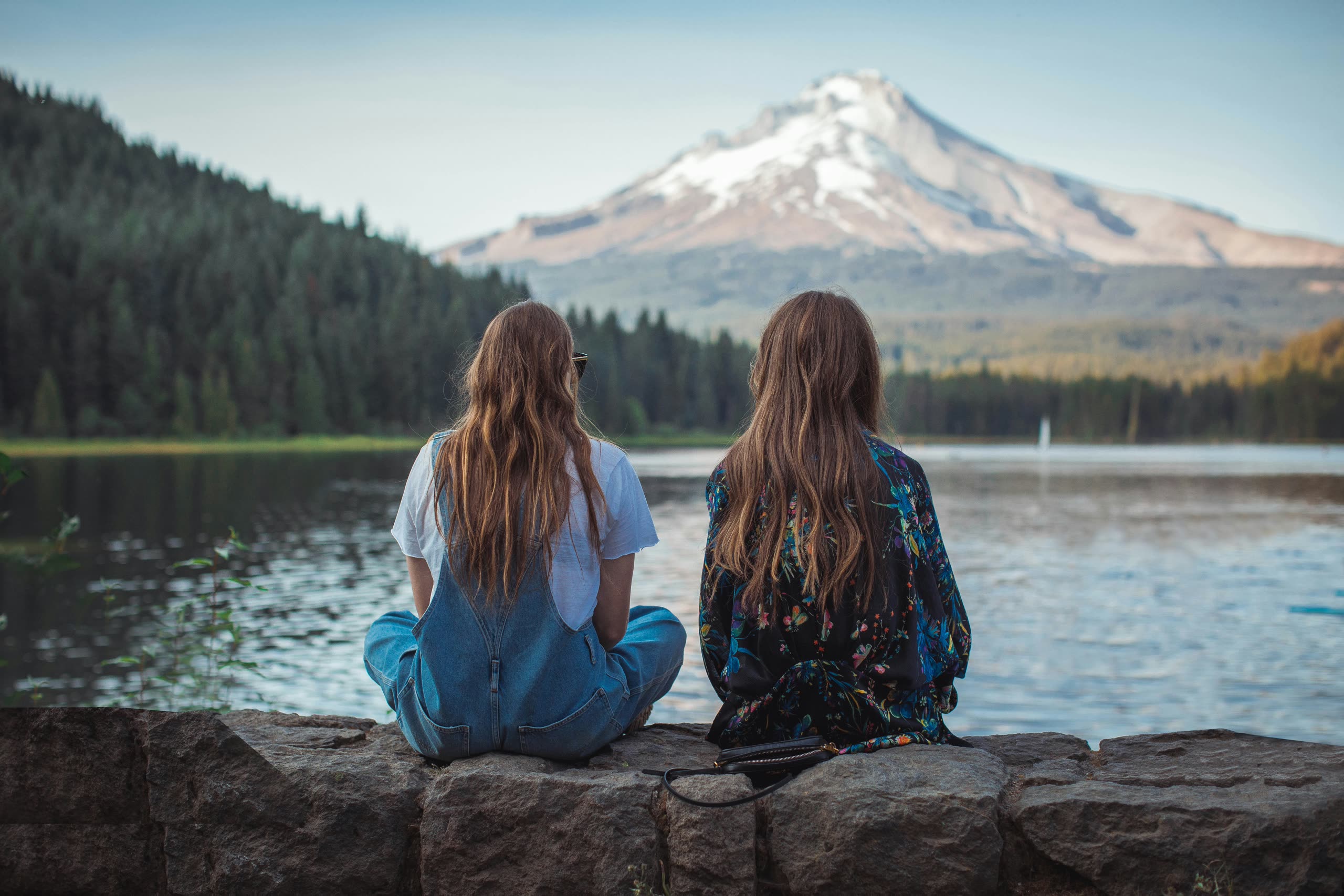 Two friends sitting on a stone wall beside a still lake at dusk, looking out at a snow-capped mountain